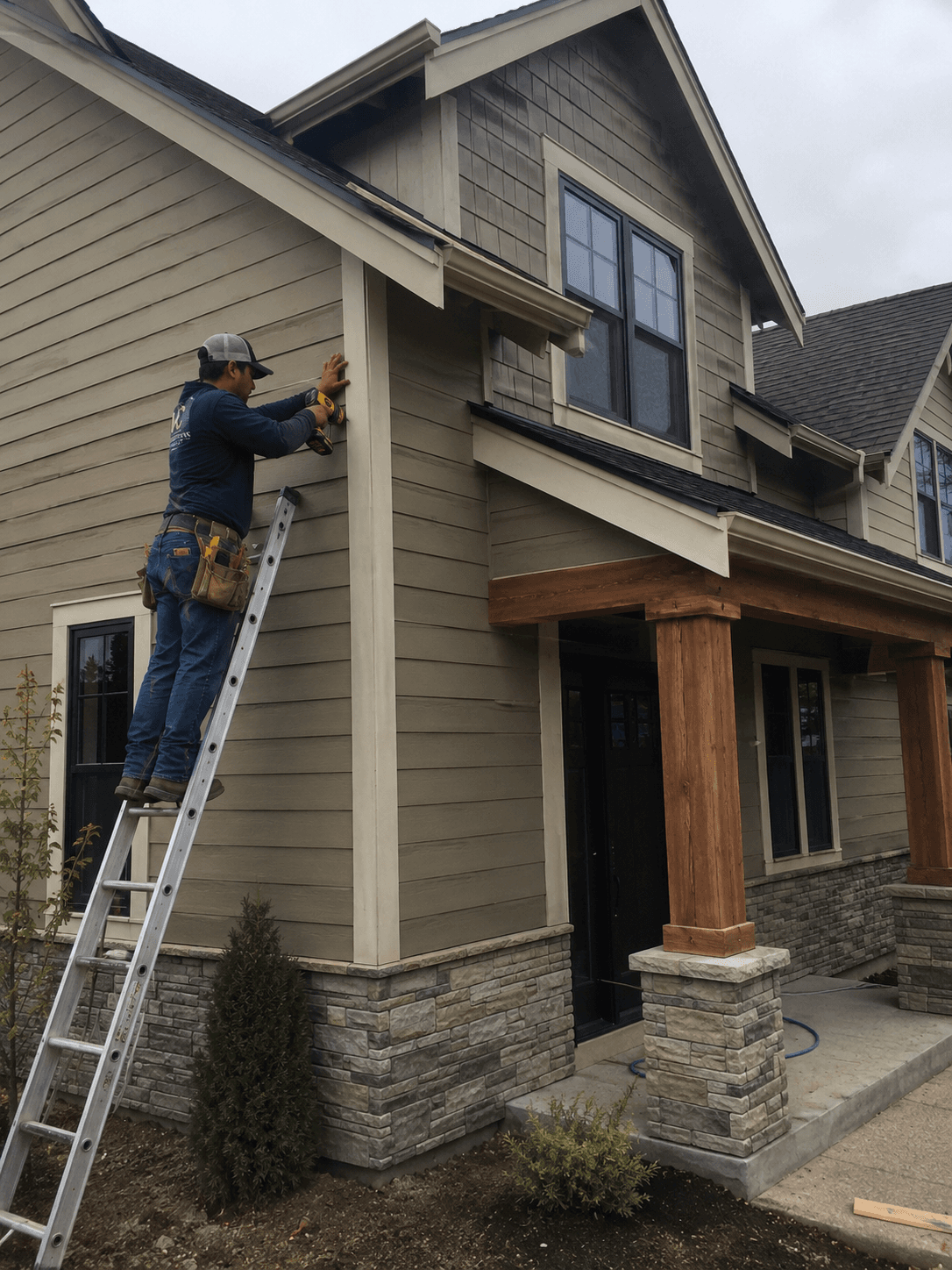 Worker on a ladder uses a power drill to install siding on a modern house.